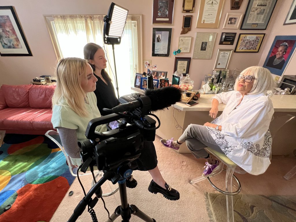 Two students interview Barbara Boyd in her home. There are items framed on the wall. Video camera and lighting are pointed at Barbara.